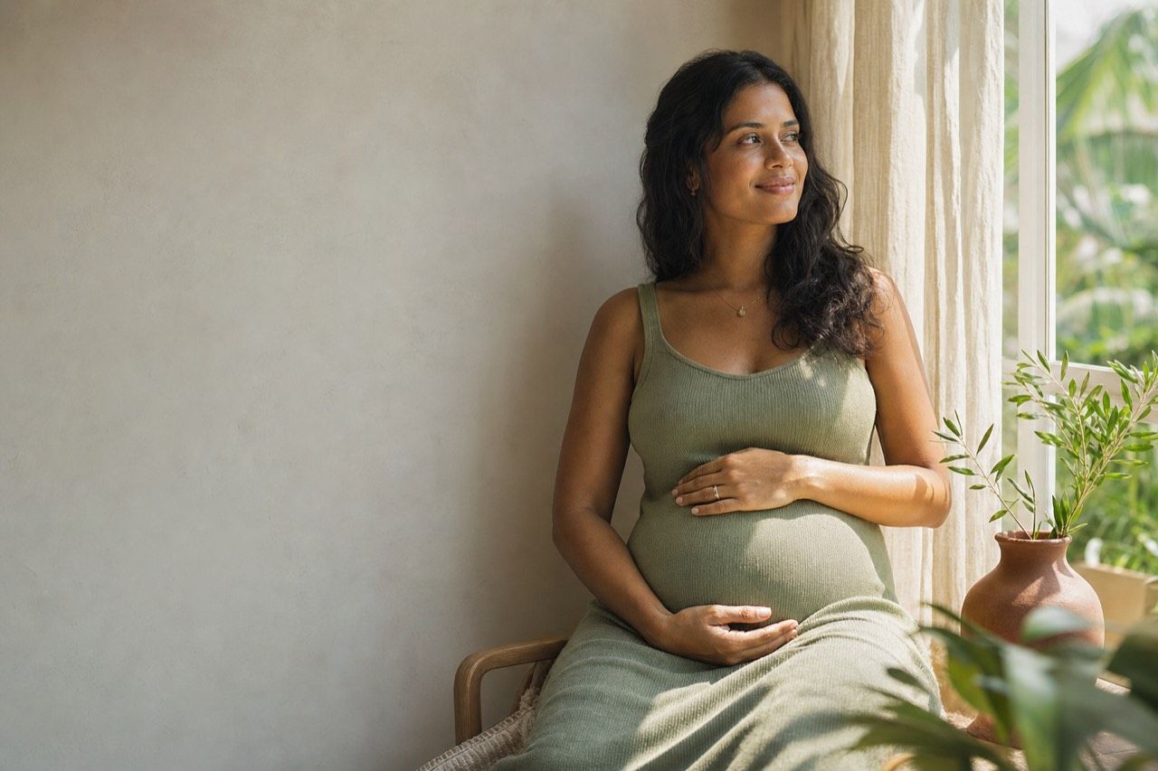Gestante sentada junto à janela com as mãos na barriga, serena, em casa acolhedora de tons claros