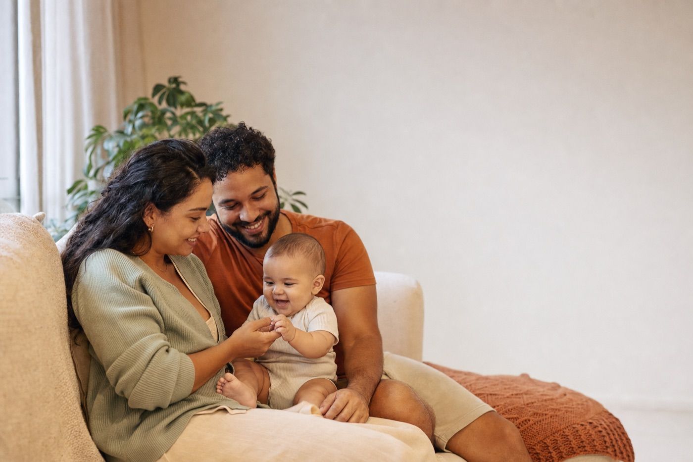 Mãe e pai sentados no sofá de casa brincando com o bebê no colo, em clima acolhedor