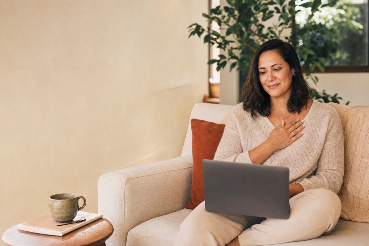 Mulher no sofá de casa em sessão de terapia online pelo notebook, com caderno e chá ao lado